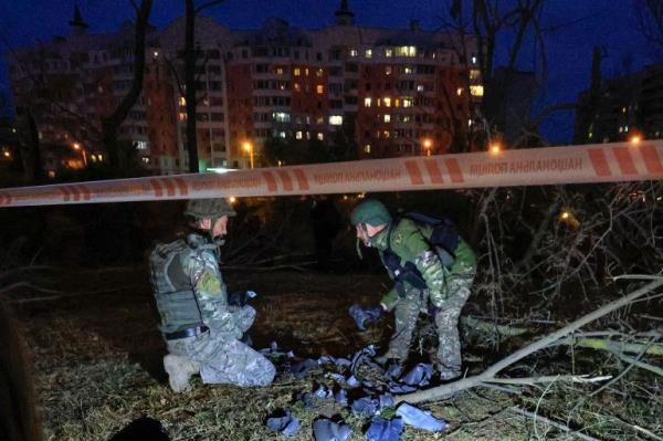 Experts collecting weapons' debris at the scene of the latest Kharkiv attack. They are in uniforms and one is kneeling on the ground while another is bending over to look at something. They are working behind a police tape. It is dark. There is an apartment block behind them with some lights on.