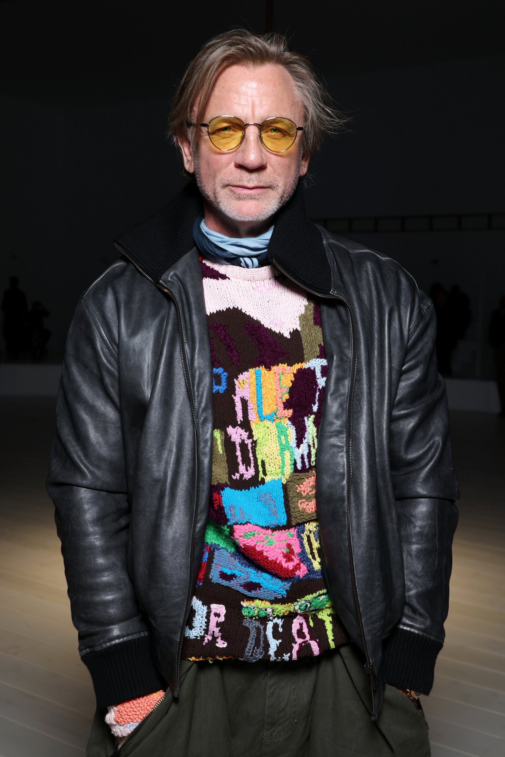 Daniel Craig poses at the Loewe Paris Womenswear Spring-Summer 2025 in a bomber jacket and unusual pink and dark red patterned jumper